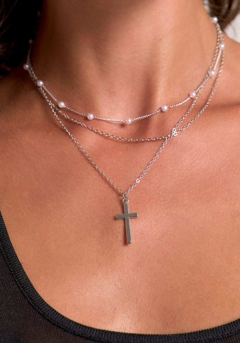A close-up of a tan, smooth neck wearing a Princess Goes Hollywood dreischichtige Silberoptik-Halskette: one with pearls, one plain chain, and one with a minimalist Kreuzanhänger, styled with a black tank top.