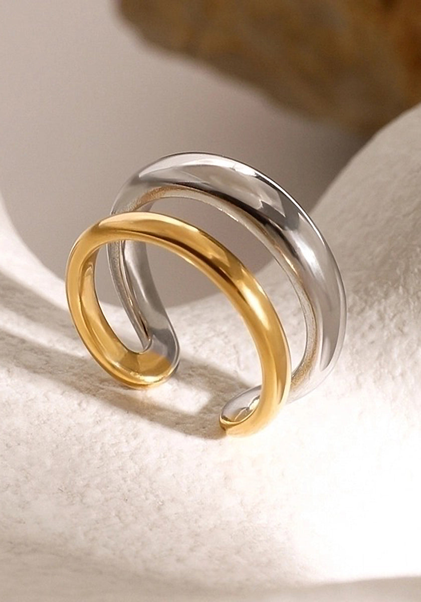 A close-up of a Princess Goes Hollywood zweifarbig Edelstahl Ring on a textured white surface. The verstellbar open ring features intertwined polished gold and silver bands in a minimalist, contemporary design.