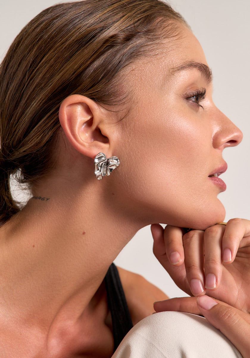 A woman with slicked-back brown hair wears große, strukturierte Edelstahl Ohrstecker from Princess Goes Hollywood. Resting her chin on her hand, she gazes right, radiant-skinned in a black top against a softly lit neutral background.