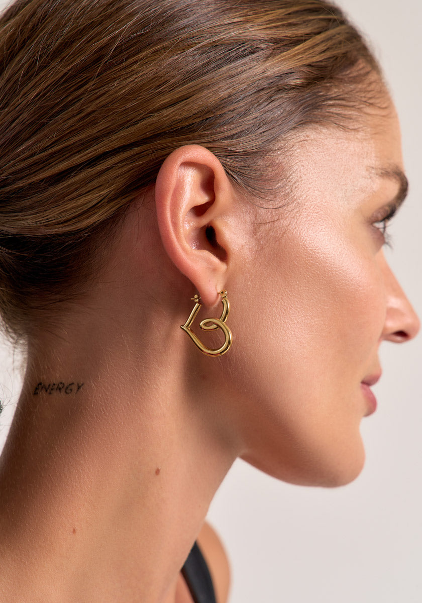 A woman with slicked-back brown hair and glowing skin wears Princess Goes Hollywood gold Herzohrringe with 18K Goldbeschichtung. The ENERGY tattoo behind her ear stands out against a plain, light background.