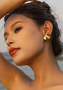A woman with dark hair pulled back poses outdoors by water, tilting her head and raising one arm. She wears bold gold earrings from Princess Goes Hollywood, with smooth skin, defined brows, long lashes, and red lipstick.