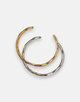 Two thin, open-ended Hoop-Armreifen aus Edelstahl by Princess Goes Hollywood are displayed on white. One has 18K Goldbeschichtung, the other is silver. They partially overlap, creating an intertwined minimalist and elegant look.