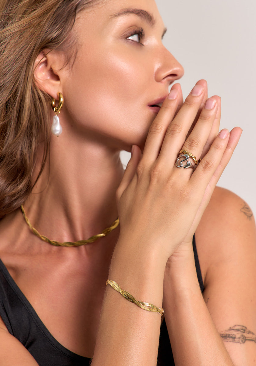 A light-skinned, brown-haired woman in profile, hands near her mouth, wears Princess Goes Hollywood’s 18K gold-plated link bracelet, matching necklace, pearl hoop earring, and a grey stone ring. Her nails are neutral; she wears a black top.