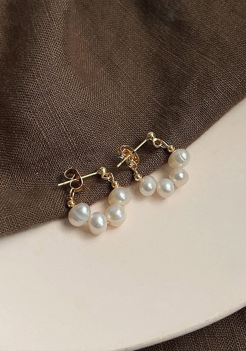 A pair of Princess Goes Hollywood gold stud earrings with three dangling Süsswasserperlen rests on soft brown fabric and a cream surface, highlighting the jewelrys elegance against the textured background.