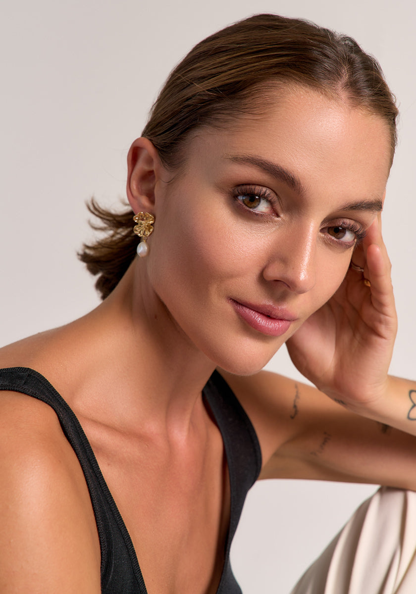 A woman with brown hair pulled back, in a sleeveless black top by Princess Goes Hollywood, smiles softly. She wears Süsswasserperle Ohrstecker, has subtle makeup, arm tattoos, and poses against a light plain background.