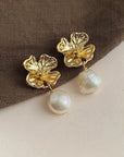 A pair of gold flower-shaped Ohrstecker by Princess Goes Hollywood, with intricate petals and sparkling centers, each featuring a dangling runde Süsswasserperle, displayed on brown textured fabric.