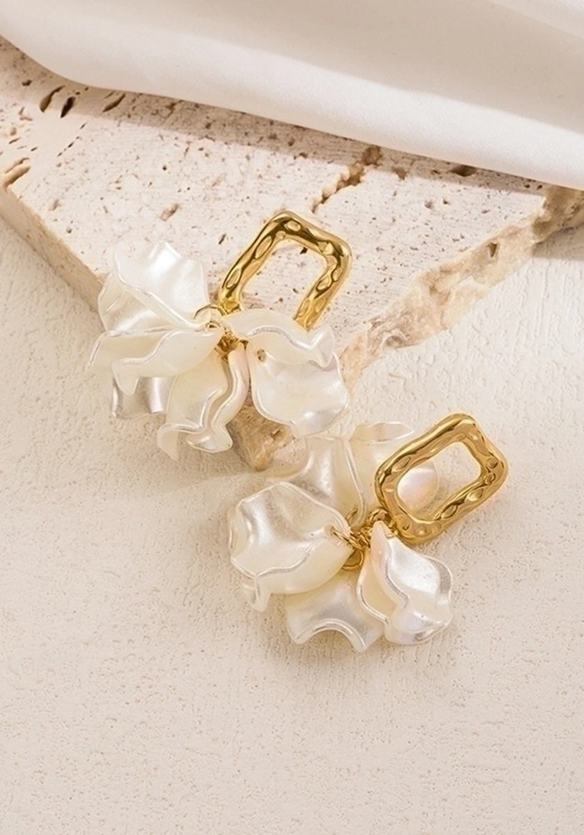 A pair of Princess Goes Hollywood gold Ohrstecker with textured rectangular Edelstahl studs and dangling wavy, iridescent white Kunstperlen, resembling flower petals, displayed on light beige stone with soft white fabric above.