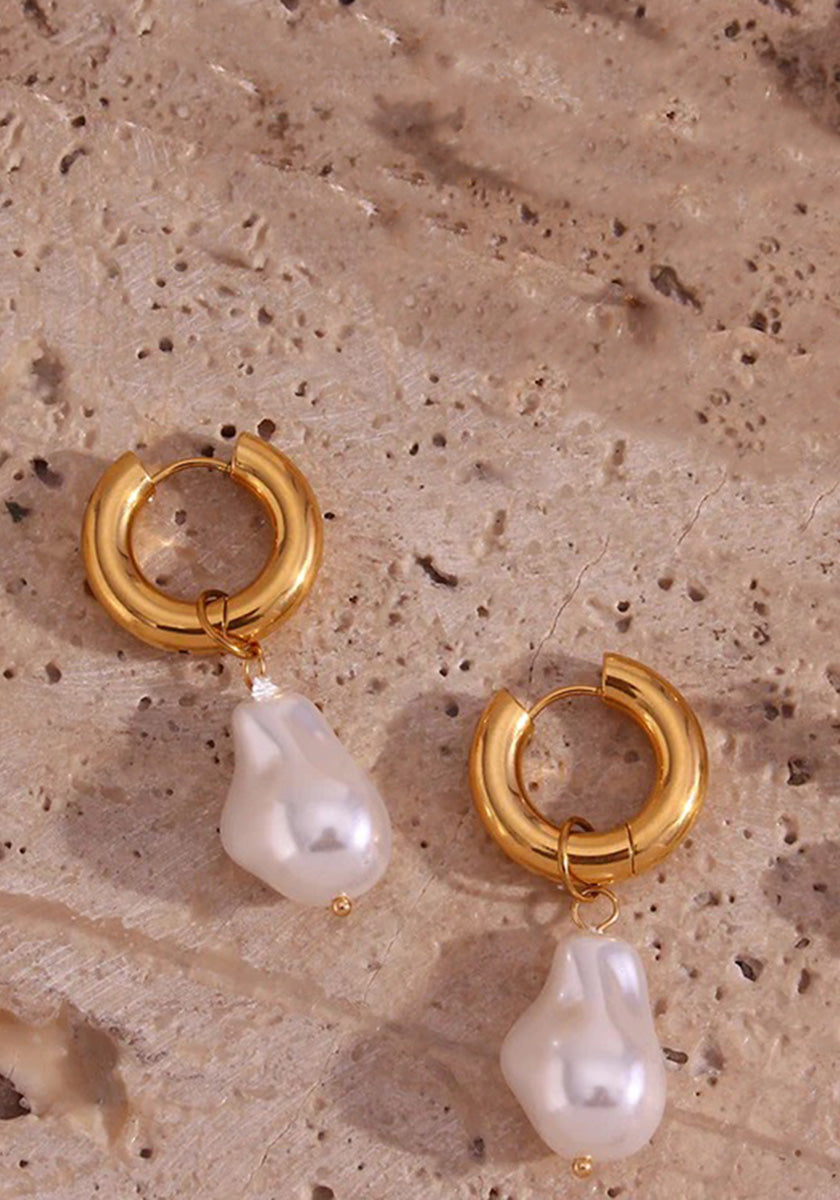 A pair of Princess Goes Hollywood Creolen Edelstahl hoop earrings with 18K Goldbeschichtung, each featuring an irregular baroque-style Süsswasserperle, displayed on a textured beige stone with natural holes and grooves.