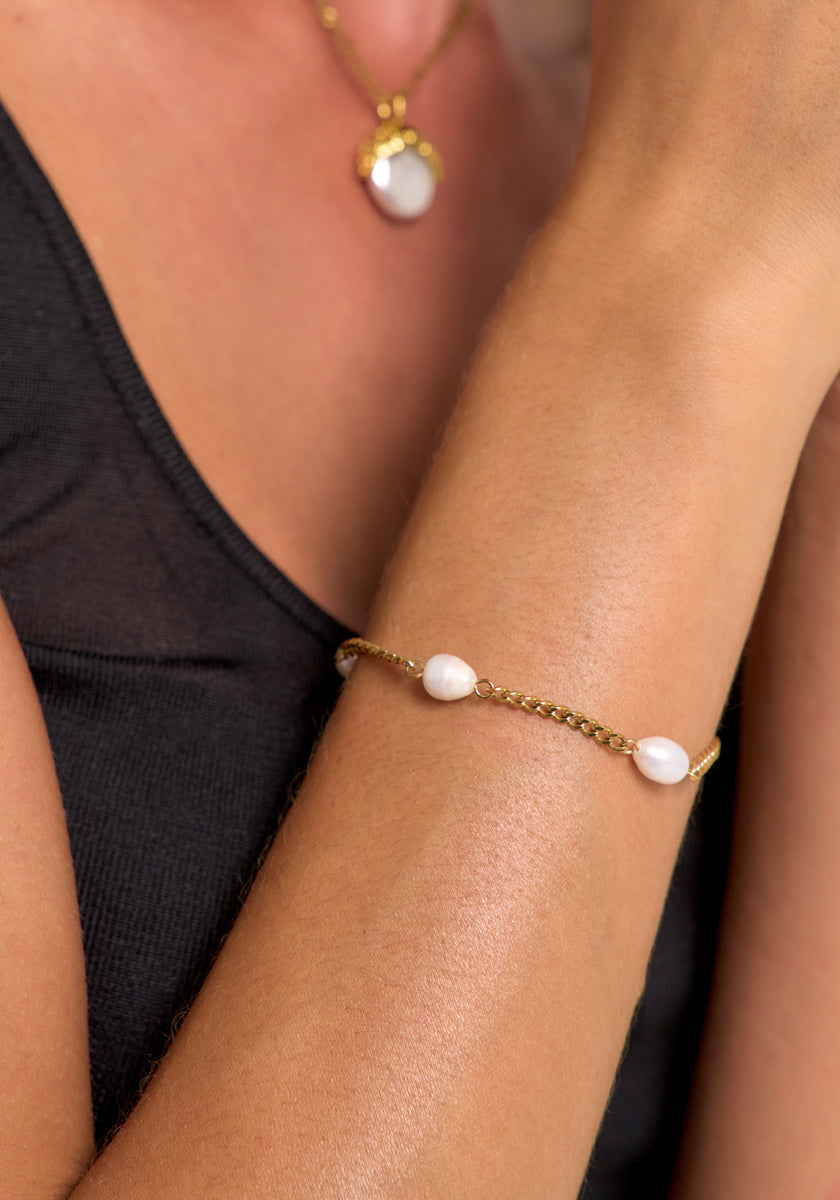 A close-up of a person with light brown skin wearing a black sleeveless top, highlighting Princess Goes Hollywood’s gold-plated bracelet with freshwater pearls and a matching gold necklace featuring a large white pearl.