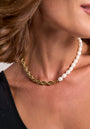 A close-up of a woman’s neck wearing a Princess Goes Hollywood necklace: half 18K Goldbeschichtung gedrehte Gliederkette, half weiße Süsswasserperlen. She has shoulder-length brown hair, a black asymmetrical top, and a neutral background.