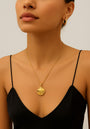 A woman in a black spaghetti strap top and gold hoop earrings showcases a Princess Goes Hollywood Gliederkette mit Muschelanhänger aus 18K-Goldbeschichtung. Neutral lips, pulled-back hair, and a light background highlight her elegant jewelry and neckline.