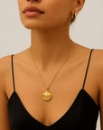A woman in a black spaghetti strap top and gold hoop earrings showcases a Princess Goes Hollywood Gliederkette mit Muschelanhänger aus 18K-Goldbeschichtung. Neutral lips, pulled-back hair, and a light background highlight her elegant jewelry and neckline.