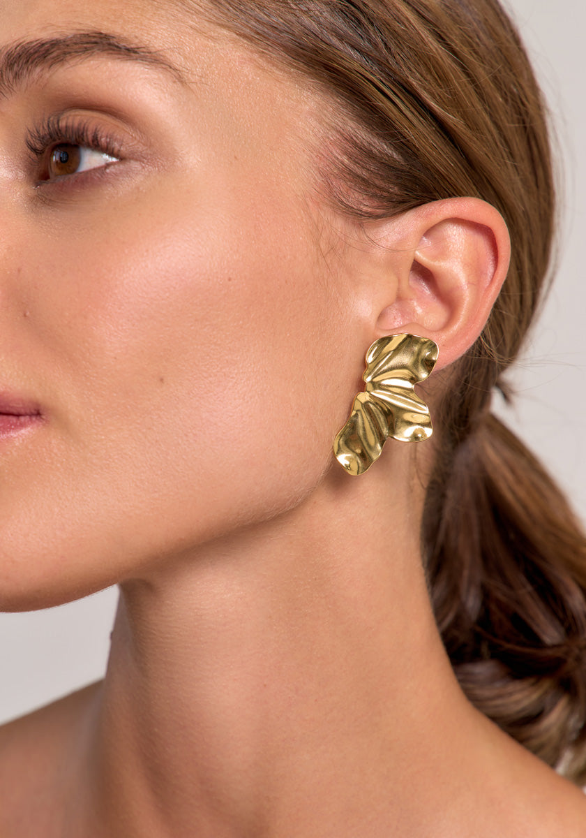 Close-up of a woman’s face and ear, highlighting a large, gold earring from Princess Goes Hollywood. Its abstract design, shiny surface, and unique folded form are accentuated by her smooth skin and low ponytail.