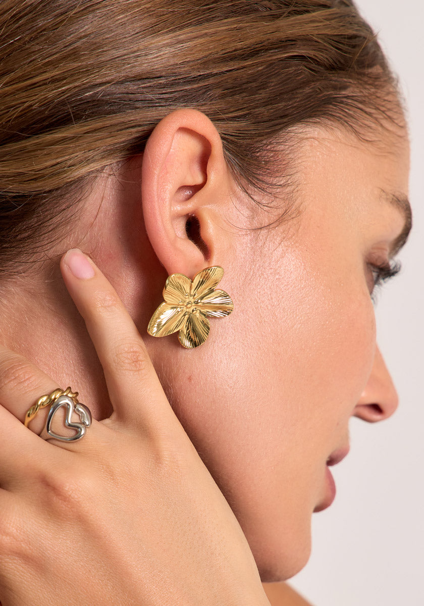 Close-up of a womans ear wearing large, gold floral stud earrings by Princess Goes Hollywood. Her brown hair is tucked back, and she gently touches her earlobe while wearing a stainless steel heart-shaped ring. The background is neutral.