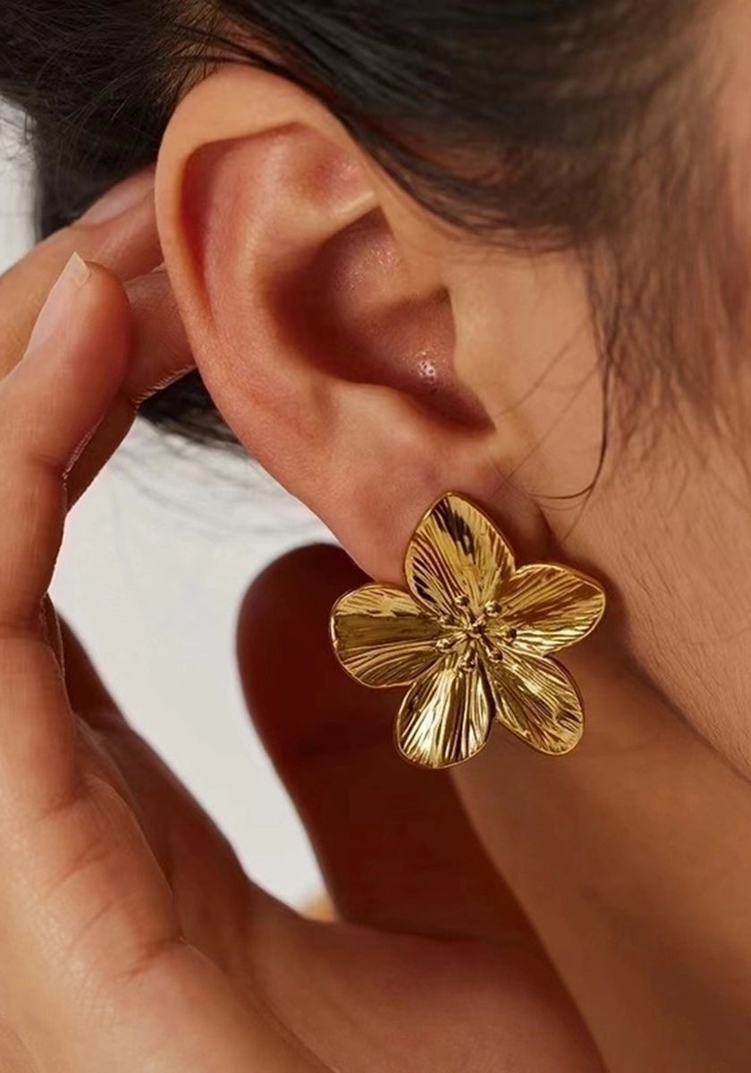 Close-up of a person’s ear adorned with gold floral stud earrings by Princess Goes Hollywood. Their hand gently touches the face, while dark hair is pulled back to highlight the shiny gold stainless steel earring.