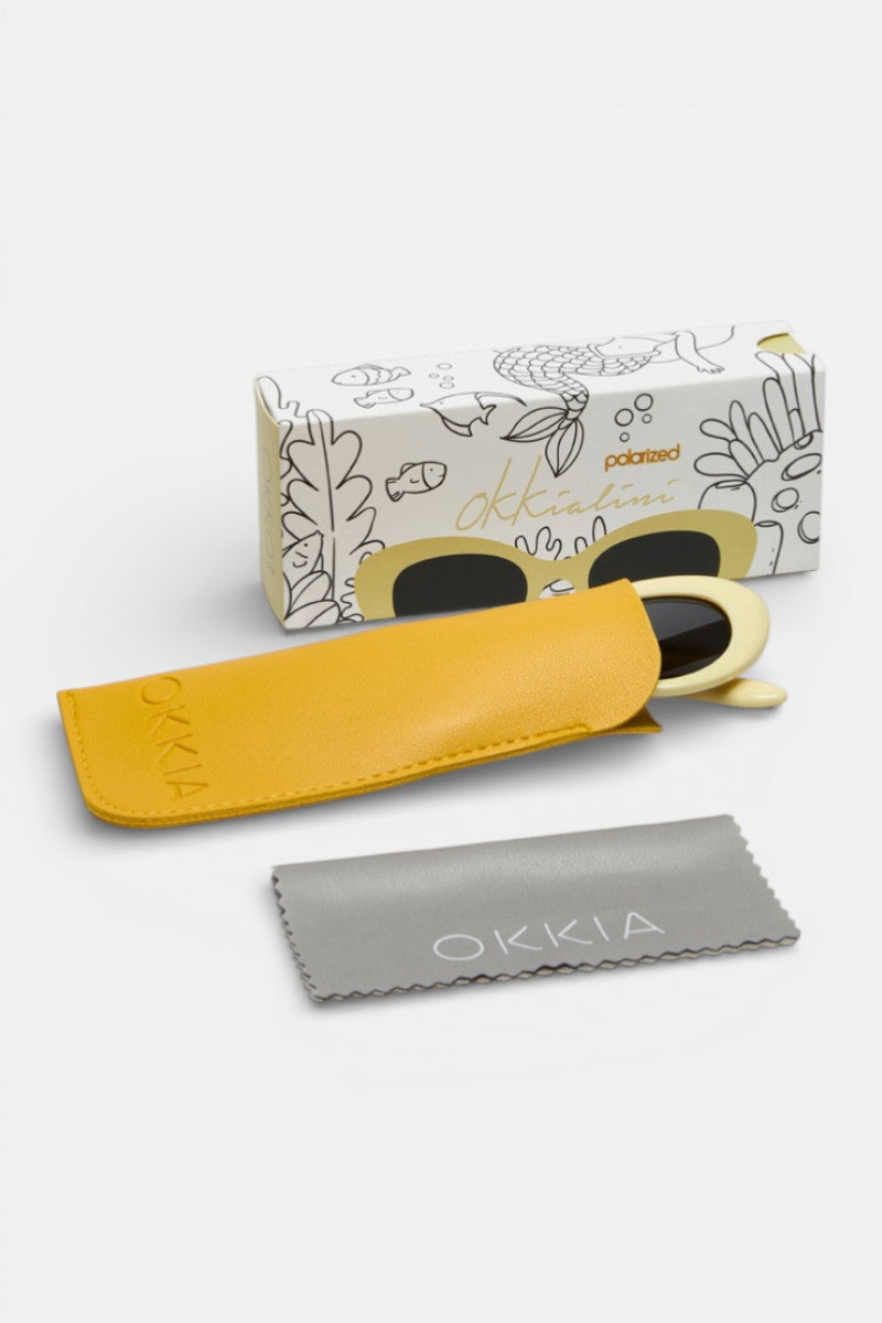 Eine Okkia-Sonnenbrille mit cremefarbenem Rahmen und dunklen Gläsern sitzt teilweise in einem gelben Etui. Auf der Vorderseite befindet sich ein graues Okkia-Putztuch und auf der Rückseite eine dekorative Okkia-Box mit verspielten, von der Natur inspirierten Linien.