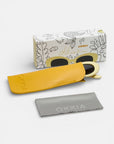 Eine Okkia-Sonnenbrille mit cremefarbenem Rahmen und dunklen Gläsern sitzt teilweise in einem gelben Etui. Auf der Vorderseite befindet sich ein graues Okkia-Putztuch und auf der Rückseite eine dekorative Okkia-Box mit verspielten, von der Natur inspirierten Linien.