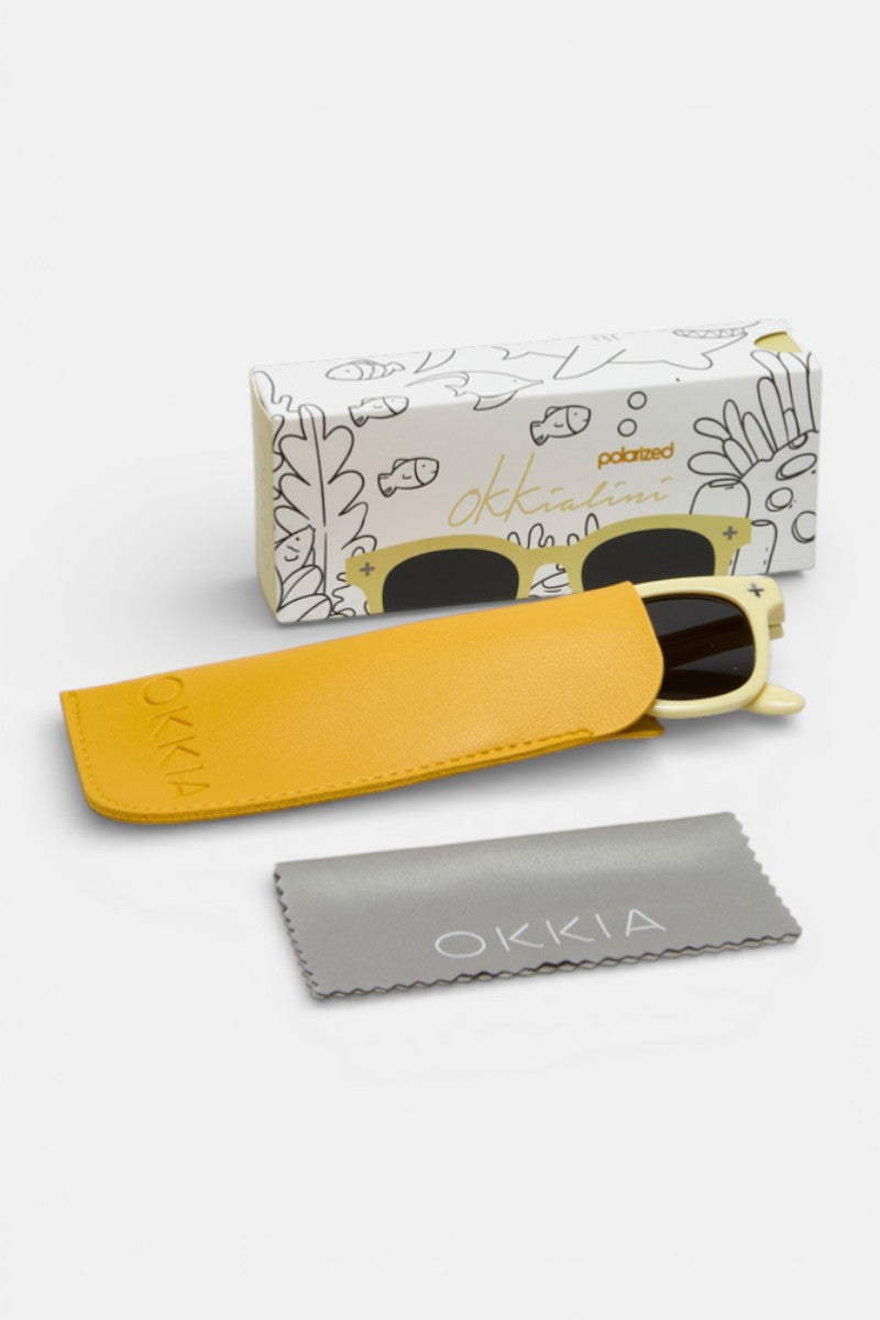 Eine Okkia Cat-Eye-Sonnenbrille mit cremefarbenem Rahmen aus der Okkialini-Kollektion liegt teilweise in einem gelben Okkia-Etui. Davor liegt ein graues Tuch, dahinter eine weiße Schachtel mit verspielten Zeichnungen und der Aufschrift "Okkiaine polarisierte Kinder Sonnenbrille".