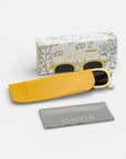 Eine Okkia Cat-Eye-Sonnenbrille mit cremefarbenem Rahmen aus der Okkialini-Kollektion liegt teilweise in einem gelben Okkia-Etui. Davor liegt ein graues Tuch, dahinter eine weiße Schachtel mit verspielten Zeichnungen und der Aufschrift "Okkiaine polarisierte Kinder Sonnenbrille".