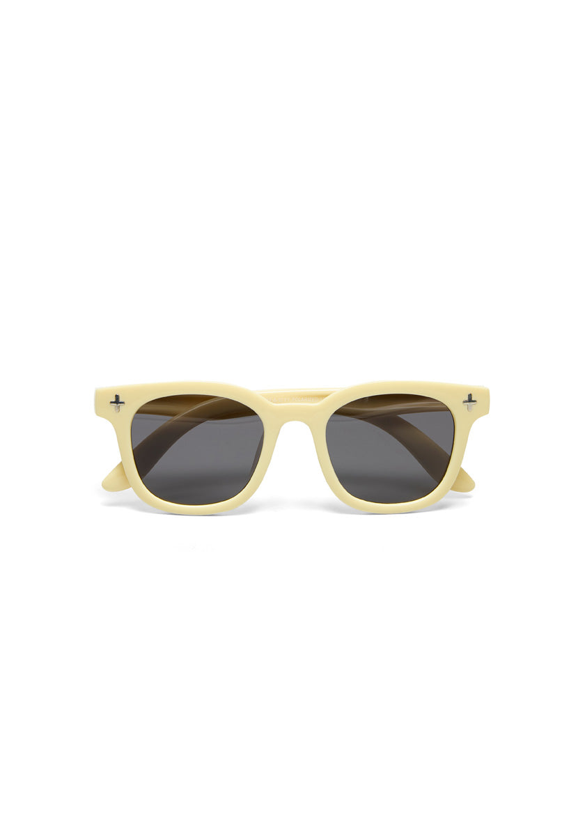 Front view of an Okkia Kinder-Sonnenbrille with thick pale yellow frames, dark oval lenses, and small black cross-shaped details. Made from flexible TPEE material for reliable protection. Shown on a plain white background.