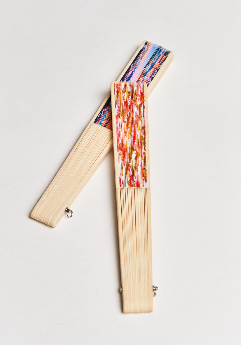 Two closed Handfächer Miami fans by Princess Goes Hollywood, made of light wood, are crossed in an X shape. Their tops display colorful abstract Miami-Prints: one with vertical red and orange strokes, the other with multicolored lines.