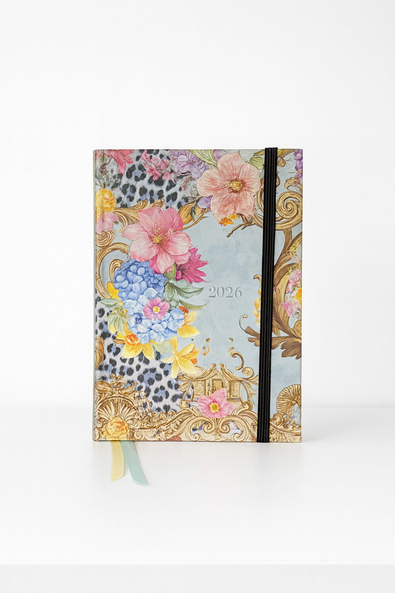 A 2026 Jahresplaner by Princess Goes Hollywood stands upright against a white background. Its cover displays pink, yellow, and blue flowers, gold ornate details, subtle leopard print, plus an elastic band and two pastel bookmarks.