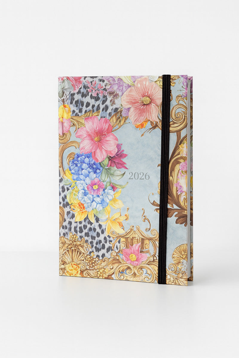 A decorative Princess Goes Hollywood Jahresplaner 2026 stands upright against a white background, featuring colorful flowers, gold baroque patterns, a subtle leopard print, and a black elastic band closure along the right edge.