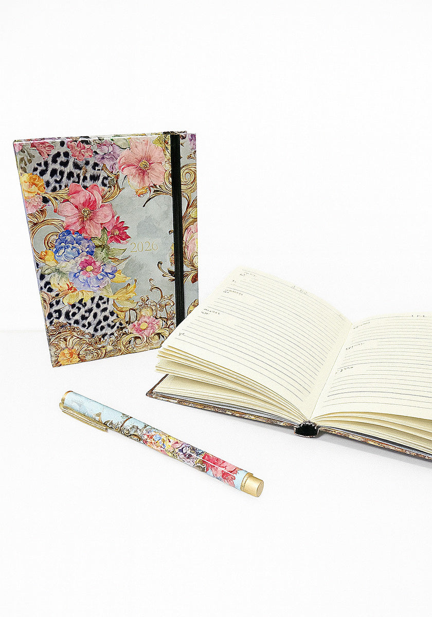 A vibrant floral Princess Goes Hollywood Terminplaner 2026 stands upright beside an open lined notebook and matching pen, all set against a white background with pink, purple, blue flowers and black-white leopard print accents.