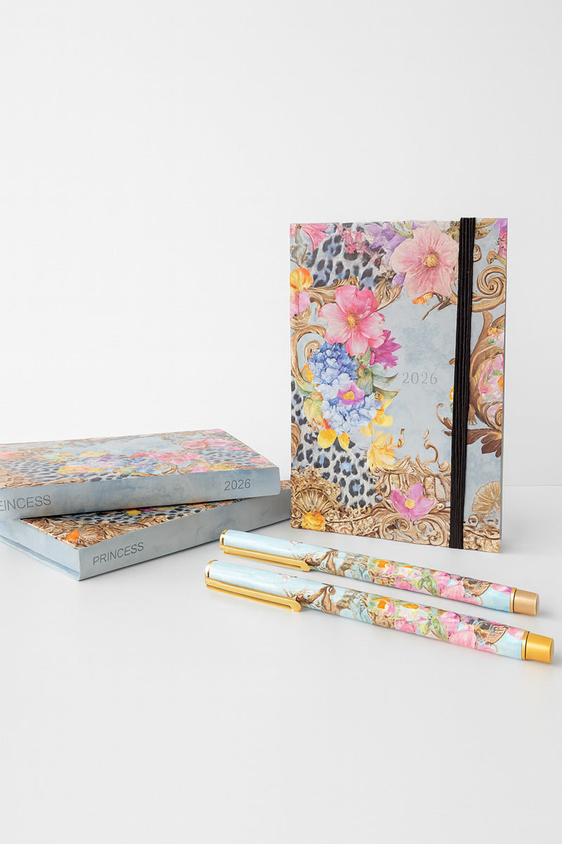 A Princess Goes Hollywood floral Terminplaner 2026 with gold and animal print stands upright by its matching box, while another box lies flat. Two stylish Kugelschreiber with blue, gold, and floral designs rest in front in an elegant, bright scene.