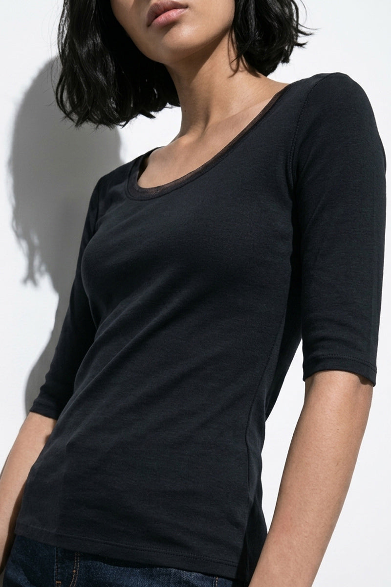 Eine Frau mit kinnlangem schwarzen Haar trägt ein bequemes, schwarzes 3/4-Arm Baumwollshirt von Princess Goes Hollywood und dunkle Jeans. Sie steht vor weißem Hintergrund, teils im Schatten, das Gesicht leicht seitlich mit geschlossenen Lippen.