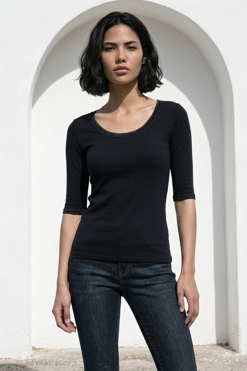 Eine Frau mit kurzen dunklen Haaren, die ein Princess Goes Hollywood Baumwolle Shirt mit 3/4-Arm und dunkler Jeans trägt, steht vor einer weißen Bogenwand im Sonnenlicht und blickt mit neutralem Gesichtsausdruck in die Kamera.