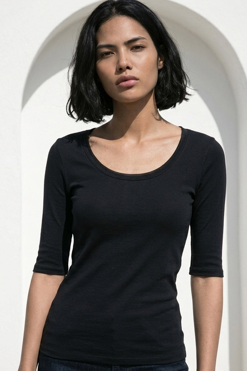 Eine Frau mit mittlerem Hautton und schulterlangem schwarzem Haar steht vor einem weißen, gewölbten Hintergrund und trägt ein 3/4-Arm-Baumwollhemd von Princess Goes Hollywood. Sie schaut mit neutralem Blick direkt in die Kamera.