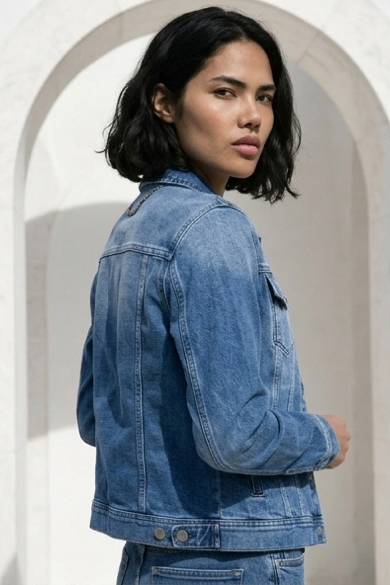 Eine Person mit schulterlangem schwarzem Haar, die ein blaues Damen-Hemd im Jeans-Stil von Princess Goes Hollywood trägt, steht im Freien vor einem weißen Steinbogen und blickt ruhig über ihre linke Schulter.