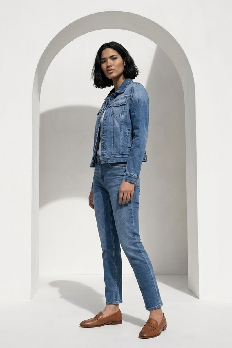 Eine Frau mit schwarzem Kurzhaarschnitt steht seitlich unter einem weißen Bogen. Sie trägt eine hellblaue Princess Goes Hollywood Damen Hemd aus Denim, passende Jeans, ein dunkles Top und braune Slipper in einer minimalistischen Umgebung mit weißen Wänden und Schatten.