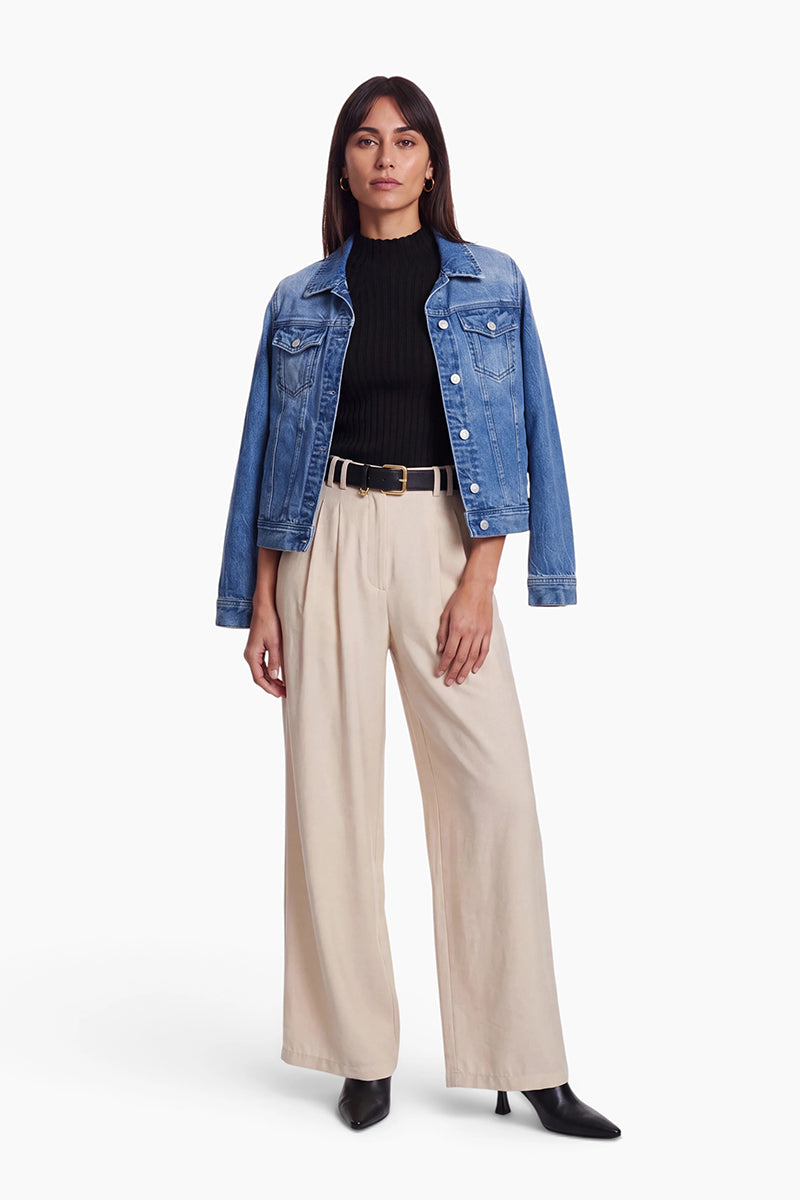 Eine Frau trägt eine blaue Jeansjacke mit klassischem Hemdkragen und Brusttaschen über einem schwarzen Top mit Stehkragen und einer beigen Hose mit weitem Bein. Dazu trägt sie einen schwarzen Gürtel mit goldener Schnalle und schwarze Stiefel mit spitzer Zehenpartie.
