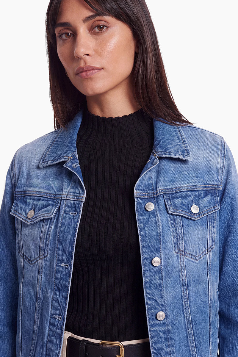 Eine Frau trägt eine hellblaue Jeansjacke mit Brusttaschen über einem schwarzen, gerippten Oberteil mit Stehkragen. Die Jacke ist offen, sodass ein schwarzer Gürtel mit goldener Schnalle an ihrer Taille zu sehen ist.