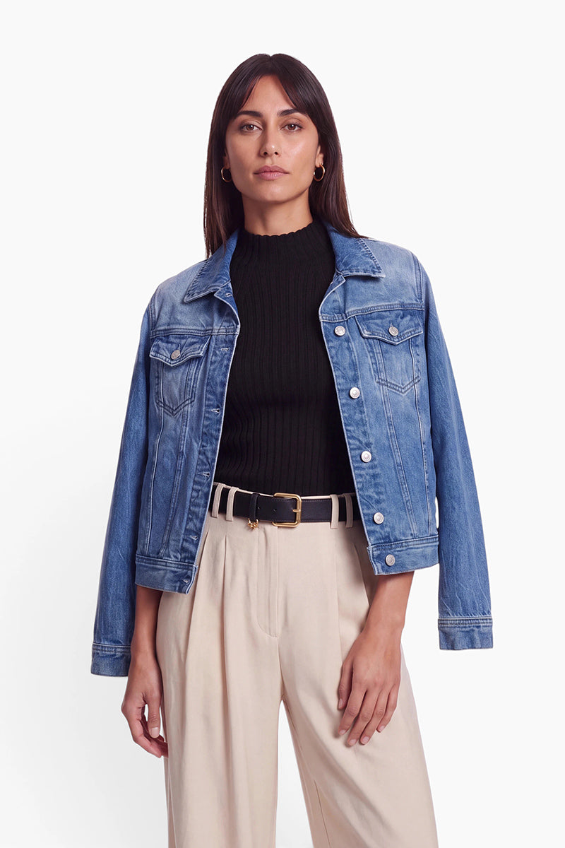 Eine Frau trägt eine blaue Jeansjacke mit klassischem Hemdkragen über einem schwarzen, gerippten Rollkragenpullover. Dazu trägt sie eine helle Hose, die mit einem schwarzen Gürtel mit goldener Schnalle zusammengehalten wird.
