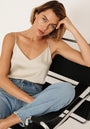 A woman with short, light brown hair sits on a black modern chair, wearing a cream-colored Princess Goes Hollywood Camisole Top with adjustable straps and light blue jeans. She rests her head thoughtfully on her hand and gazes at the camera.