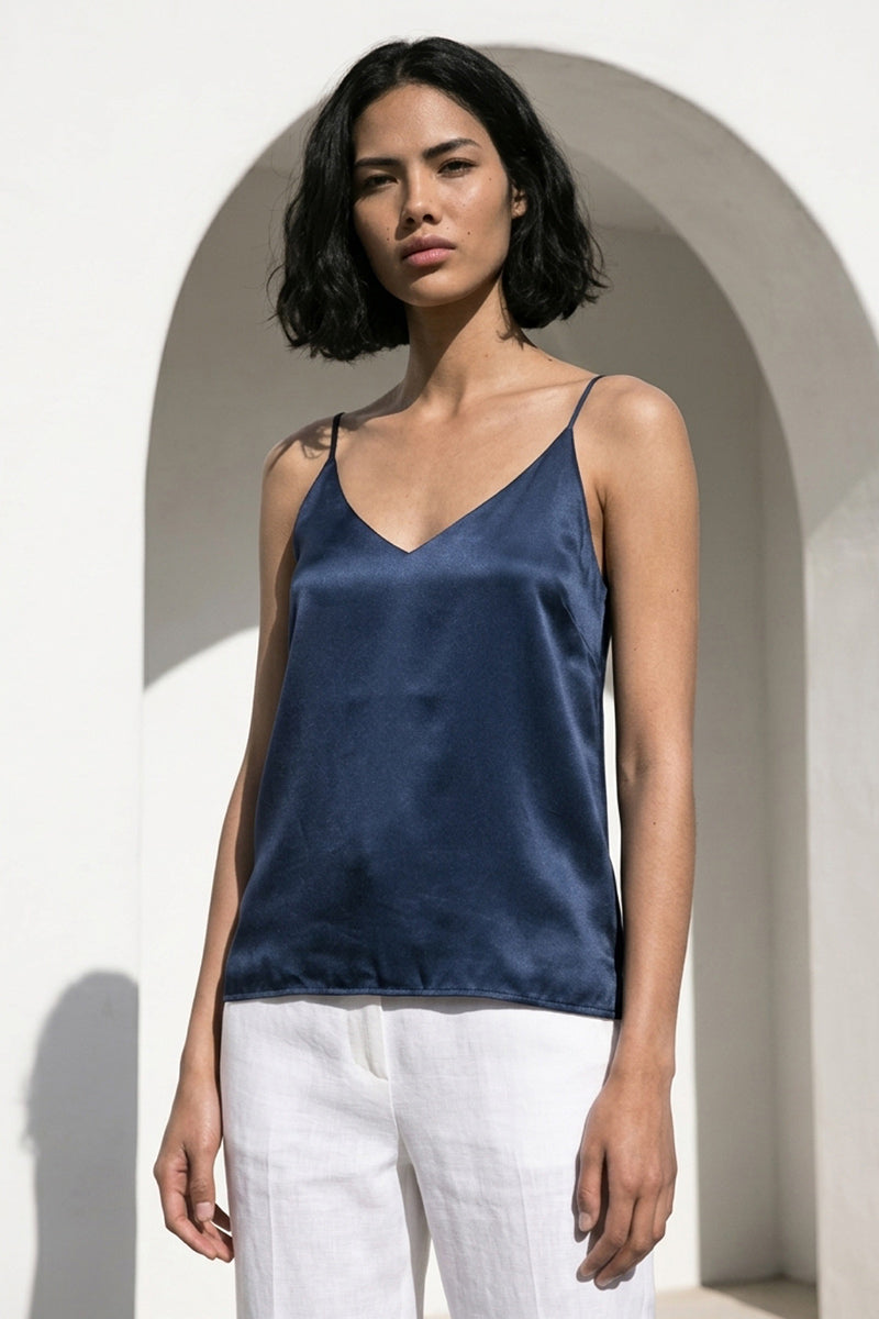 Eine Frau mit kurzen schwarzen Haaren steht vor einer weißen gewölbten Wand und trägt ein marineblaues Princess Goes Hollywood Seide Camisole Top mit V-Ausschnitt und verstellbarem Träger, gepaart mit einer weißen Hose. Sie blickt in die Kamera, die Arme entspannt an den Seiten.