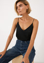 A woman with short blonde hair wears a black Princess Goes Hollywood V-Ausschnitt Camisole Top Seide with adjustable straps and high-waisted blue jeans, sitting on a light wooden bench against a plain white background, gazing slightly upward.