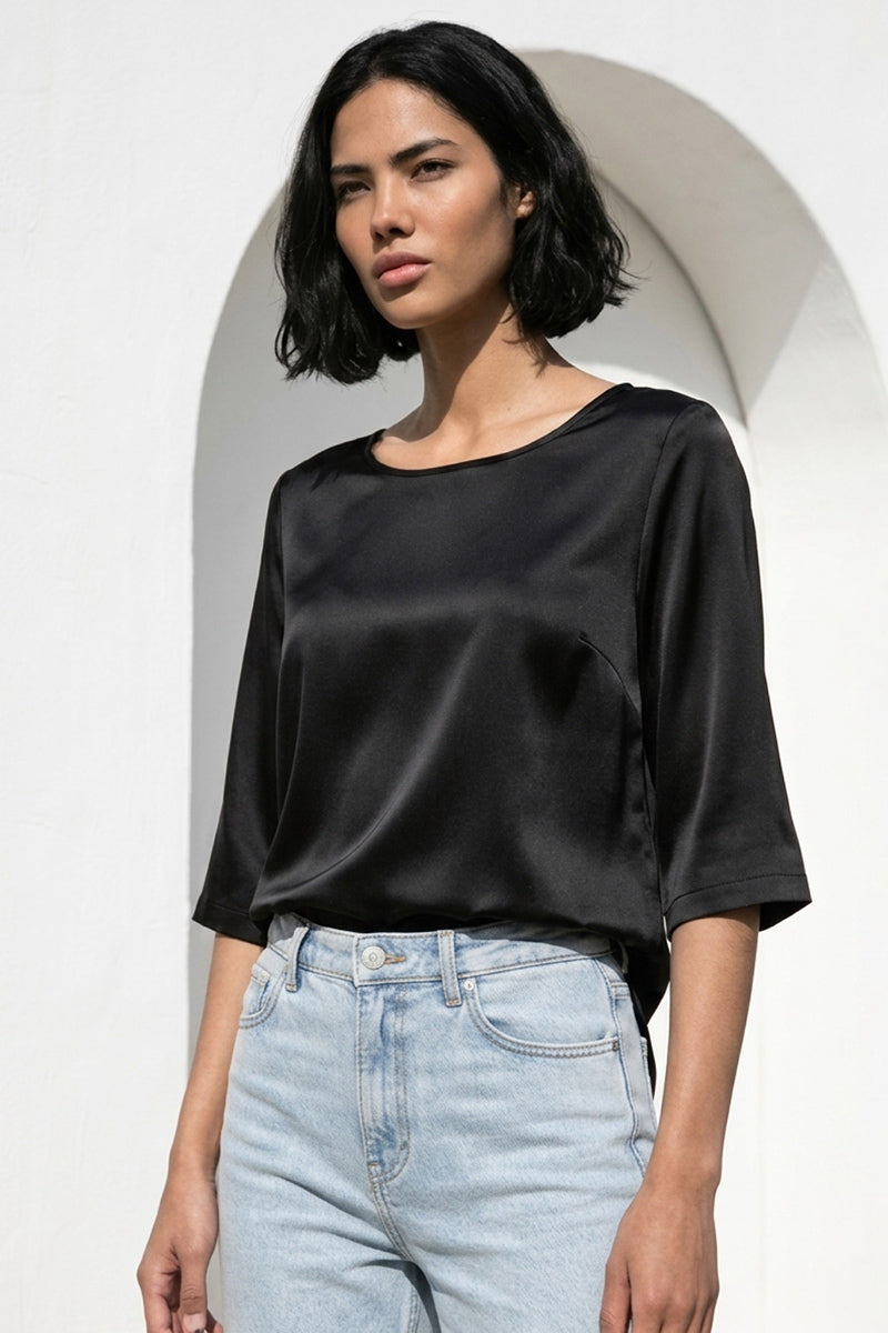 Eine Frau mit kinnlangen schwarzen Haaren trägt eine Princess Goes Hollywood Materialmix Seide Bluse mit 3/4 Ärmeln und hellblaue Jeans mit hoher Taille. Sie steht an einer weißen Wand mit einer gewölbten Öffnung und blickt mit einem neutralen Blick zur Seite.