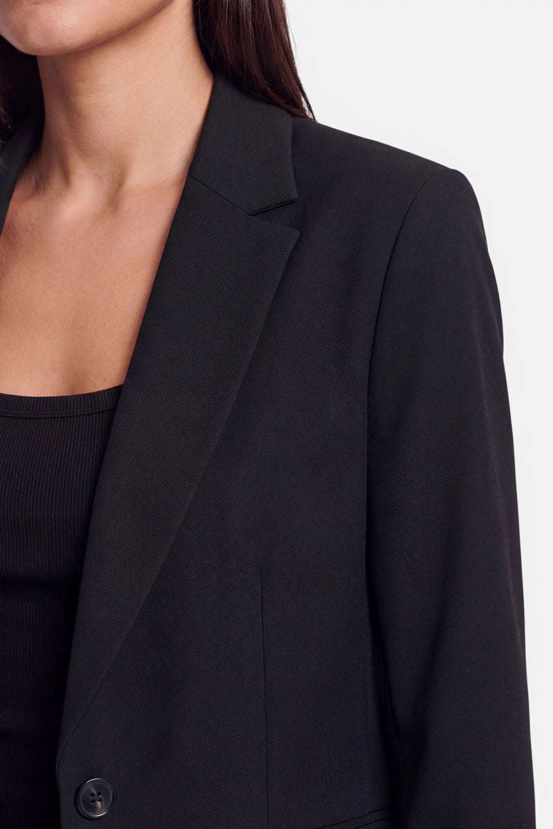 Eine Nahaufnahme einer Frau, die einen figurbetonten Blazer aus dehnbarem Webstoff über einem schwarzen, gerippten Tanktop trägt. Zu sehen sind nur ihr unterer Gesichtsbereich, ihr Hals, ihre Schulter und ein Teil ihrer Brust; ihr glattes, dunkles Haar fällt ihr über die Schulter. Der Hintergrund ist einfarbig hellgrau.