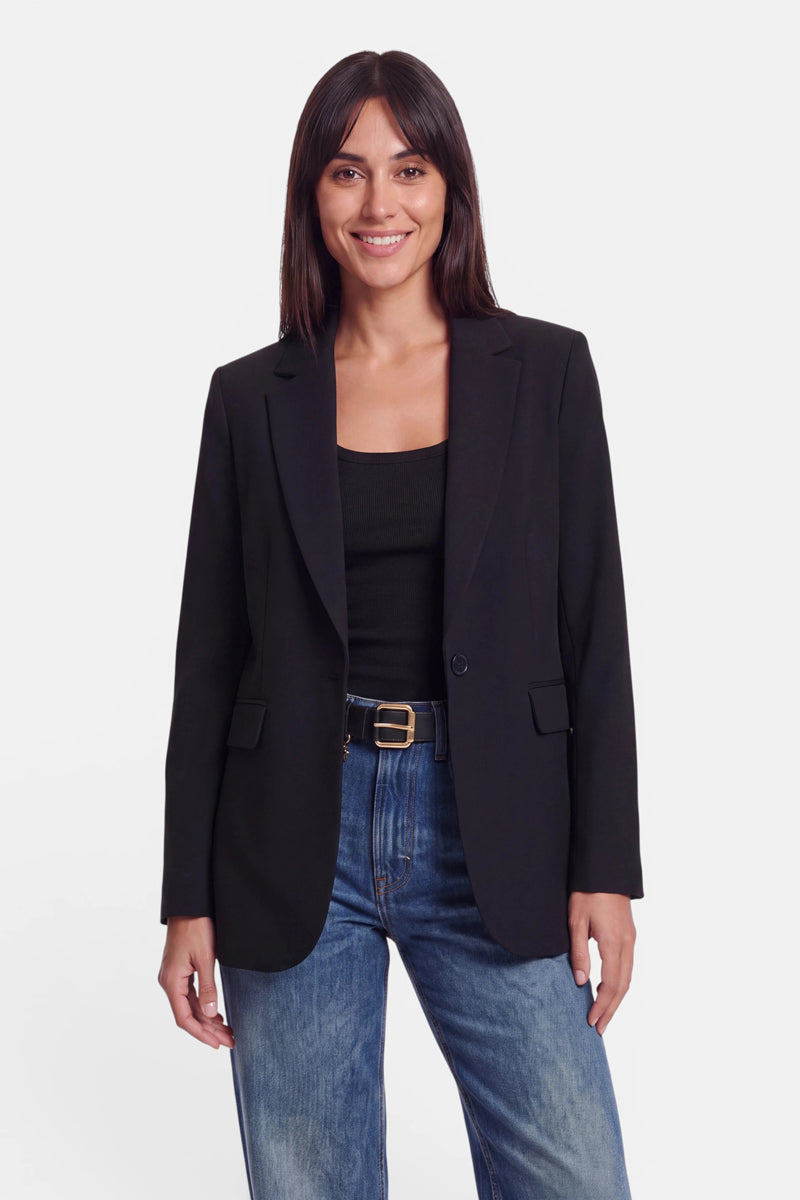Eine Frau mit glattem braunem Haar trägt einen figurbetonten Blazer aus dehnbarem Webstoff über einem schwarzen Oberteil, einer blauen Jeans und einem schwarzen Gürtel mit goldener Schnalle. Sie steht lächelnd vor einem schlichten weißen Hintergrund und bringt dabei den schmalen Schnitt ihres Outfits zur Geltung.