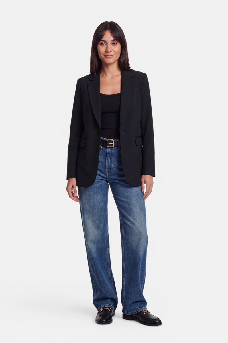 Eine Frau steht mit dem Blick nach vorne gerichtet da. Sie trägt einen figurbetonten Blazer aus dehnbarem Webstoff über einem schwarzen Oberteil, eine blaue Jeans mit weitem Bein und schwarze Loafer. Sie hat glattes, dunkles Haar mit Pony und lächelt leicht, die Hände entspannt an den Seiten, vor einem schlichten weißen Hintergrund.