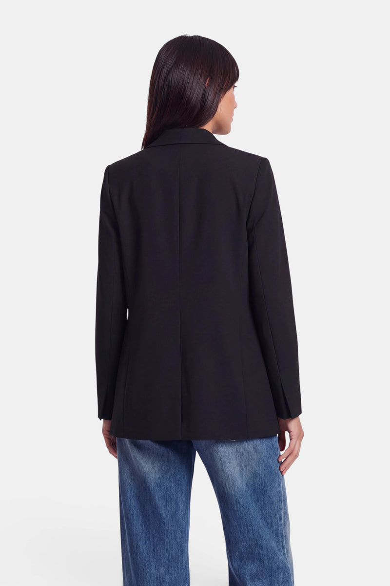 Eine Person mit langen, dunklen Haaren, von hinten gesehen, trägt einen figurbetonten Blazer in Schwarz und eine locker sitzende blaue Jeans. Der Hintergrund ist schlicht weiß, sodass die gesamte Aufmerksamkeit auf das Outfit gerichtet ist.
