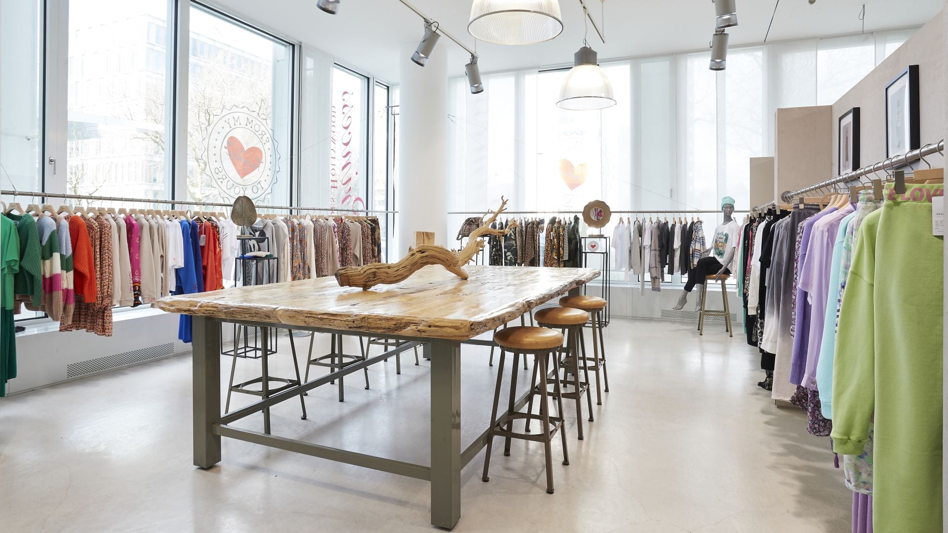A bright, modern clothing boutique with large windows, racks of colorful clothes on both sides, and a rustic wooden table with stools in the center. Natural light fills the space, and two large pendant lights hang from the ceiling above the table.