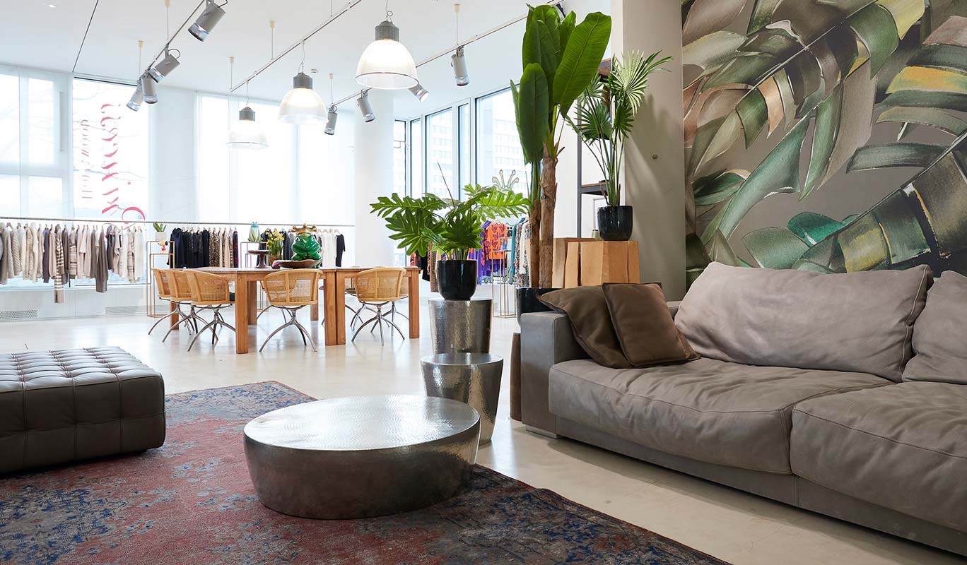 A modern boutique with a gray sofa, metallic coffee tables, potted plants, and a colorful rug in the foreground. In the background, racks of clothes, a wooden table with woven chairs, large windows, and a leafy mural are visible. The space is bright and stylish.