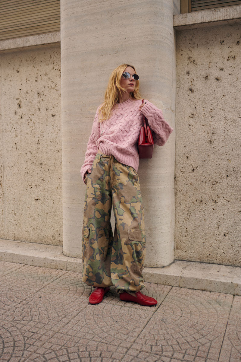 Eine blonde Frau mit dunkler Sonnenbrille lehnt an einer beigen Steinsäule. Sie trägt einen dicken, locker sitzenden rosa Zopfmusterpullover, eine übergroße Cargohose im Camouflage-Look und rote Ballerinas; über der Schulter hängt eine dunkelrote Handtasche.