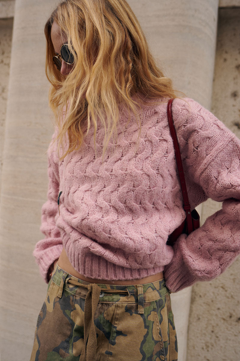 Eine Person mit welligem blondem Haar und Sonnenbrille trägt einen dicken, rosa Pullover mit Zopfmuster und eine Hose mit Camouflage-Muster. Sie steht an einer Steinsäule, neben sich eine kleine rote Umhängetasche.