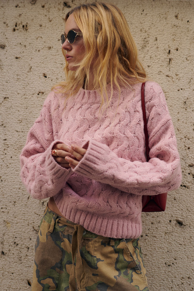 Eine Frau mit welligem blondem Haar trägt eine runde Sonnenbrille und einen dicken rosa Pullover mit Zopfmuster. Sie hat eine Camouflage-Hose an und steht vor einer strukturierten Steinmauer, wobei sie eine dunkelrote Umhängetasche bei sich trägt.